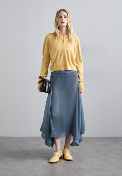 Καρντάνι μουσταρδί με υφασμάτινες κουκίδες, συνδυασμένο με μια ρέουσα μπλε πλισέ φούστα και κίτρινες παπούτσια με κοψίματα. Μικρή μαύρη τσάντα ως αξεσουάρ.