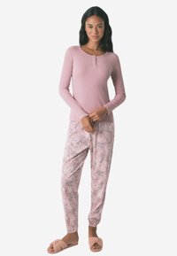 Maglione a maniche lunghe rosa chiaro con dettagli a bottoni, abbinato a pantaloni del pigiama con motivo floreale in delicate tonalità di rosa. Il modello indossa pantofole morbide.