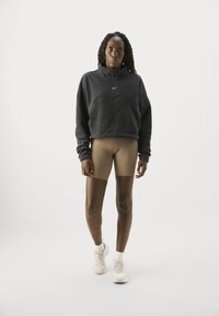 Kvinde med fletninger iført en sort pullover, brune cykelshorts, hvide sokker og lyserøde sneakers, stående foran en ensfarvet baggrund.