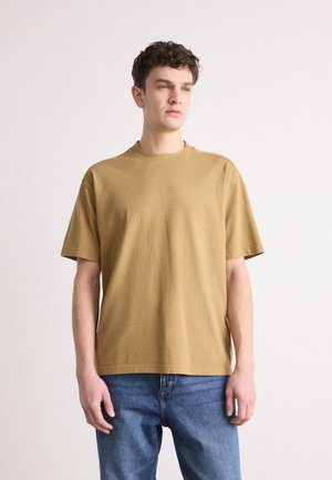 Kurzärmliges T-Shirt mit Rundhalsausschnitt aus olivgrünem Baumwollstoff. Verfügt über dezentes Branding am Halsausschnitt und einen lockeren Schnitt. Kombiniert mit blauen Jeans.
