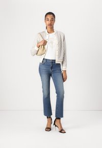 Weißes Grafik-T-Shirt unter einer gemusterten, hellen Jacke, kombiniert mit blauen Jeans und schwarzen, hochhackigen Sandalen. Hält eine kleine beigefarbene Handtasche.