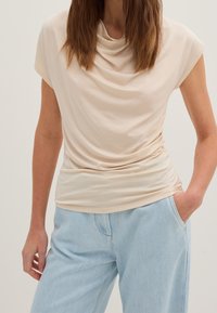 Kvinna i en krämfärgad, draperad ärmlös topp instoppad i ljusblå högmidjade jeans, med en hand i fickan och löst hår.