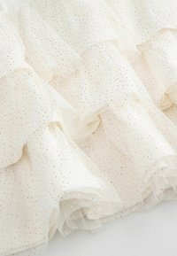 Biały tiul z warstwowymi falbanami, ozdobiony subtelnym wzorem złotych kropek. Miękka, przejrzysta tkanina z warstwowym zdobieniem na całej powierzchni.