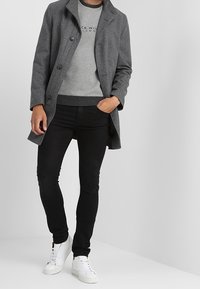 Abrigo de lana gris con solapa y botones, llevado sobre un suéter gris. Unos vaqueros negros skin fit y zapatillas blancas completan el atuendo.