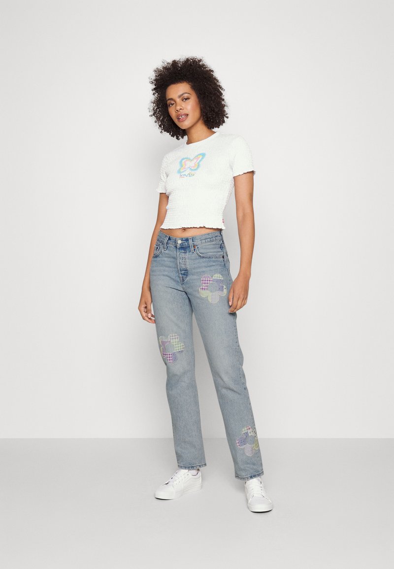 Top blanco de manga corta con tejido texturizado y un diseño colorido de mariposa, combinado con jeans azul claro que presentan parches de flores a cuadros.
