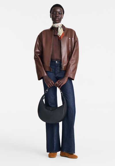 Frau trägt eine braune Lederjacke, dunkle blaue Schlagjeans, orangefarbene Schuhe, ein florales Halstuch und hält eine schwarze Halbmond-Handtasche vor einem weißen Hintergrund.