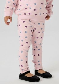 Małe dziecko ubrane w różowe legginsy i pasujący sweter z fioletowo-niebieskimi wzorami serc oraz czarne buty typu Mary Jane.