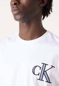 Hvid bomulds-T-shirt med et sort og lys blå broderet "ck" logo på venstre bryst og en klassisk rund halsudskæring.