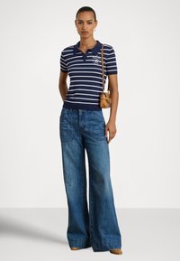 Sieviete, tērpusies tumši zils ar svītrām polokrekls, plašām zilām džinsa biksēm, ar mazu brūnu plecu somiņu un brūnām kurpēm, stāv pret vienkrāsainu fonu.