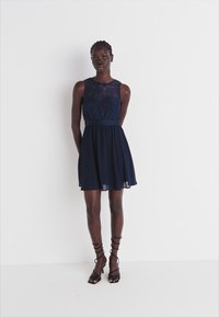 Jūras zila kleita ar mežģīņu korpusu, bez piedurknēm un pieguļošu vidukli, plūstoša svārku daļa. Saskaņota ar melniem sandaliem ar siksniņām.