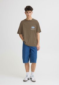 T-shirt marron oversize avec une poche poitrine et un logo, associé à un short en denim bleu longueur genou. Chaussettes blanches et baskets noires et blanches.
