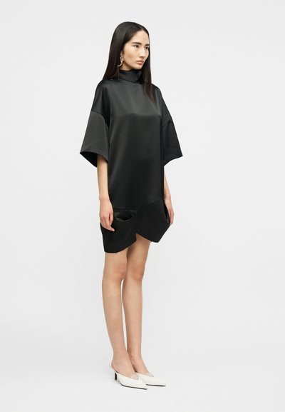 Γυναίκα που στέκεται, φορώντας ένα χαλαρό μαύρο σατέν φόρεμα με μανίκια μέχρι τον αγκώνα και λευκά παπούτσια με μυτερή μύτη, μπροστά σε απλό φόντο.