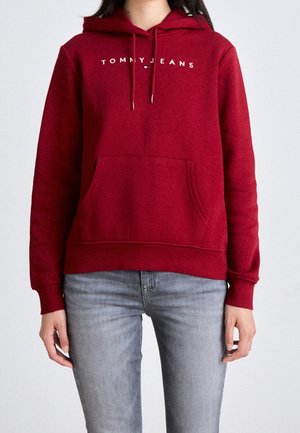 Osoba ubrana w czerwoną bluzę z kapturem z napisem "TOMMY JEANS" na piersi oraz jasnoszare dżinsy, stojąca na jednolitym tle.