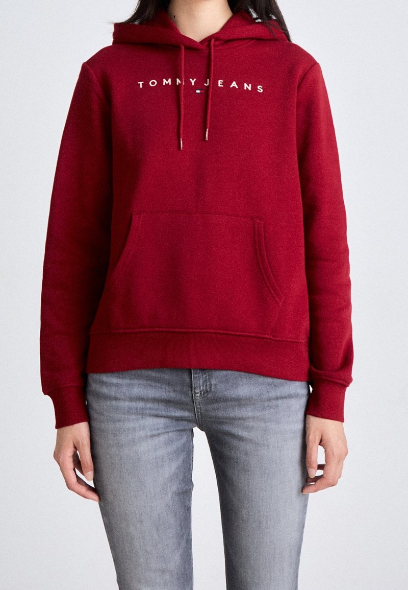 Personne portant un sweat à capuche rouge avec l'inscription « TOMMY JEANS » sur la poitrine et un jean gris clair, debout devant un fond uni.