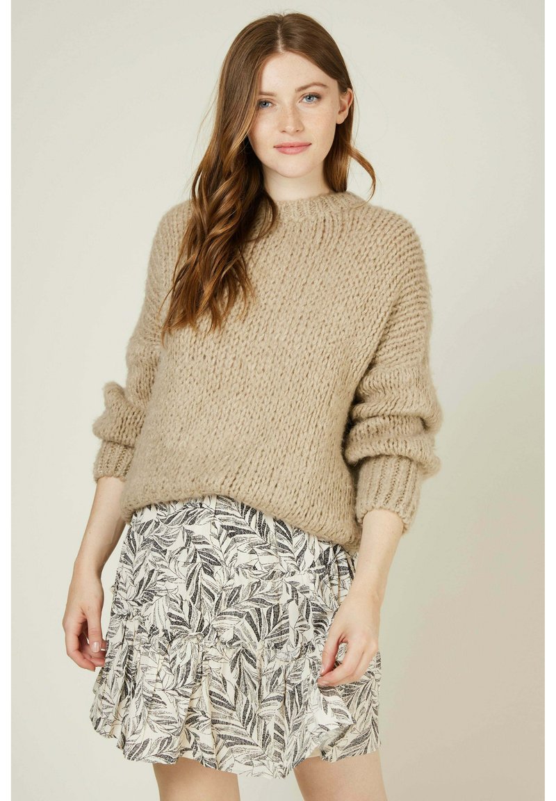 Pull en tricot épais beige avec des poignets côtelés, associé à une jupe blanche ornée de motifs de feuilles noires et d'un ourlet froncé.
