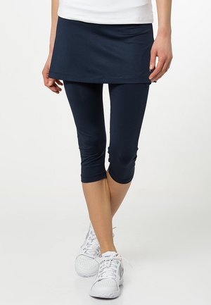 Person iført marineblå leggings med nederdel og hvide sportssko, der står med krydsede ben på en hvid baggrund.