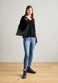 Svart ribbet genser, lyseblå skinny jeans og svarte snøresko. Modellen bærer en svart tote bag med minimalistisk design. Bakgrunn av tregulv.