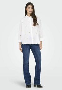 Camicia bianca con bottoni, con accenti ricamati gialli e verdi, abbinata a jeans a zampa d'elefante blu scuro e stivaletti neri alla caviglia.