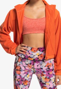 Giacca in pile arancione con polsini elastici, abbinata a un reggiseno sportivo rosa e leggings a fantasia floreale multicolore con dettaglio zip.
