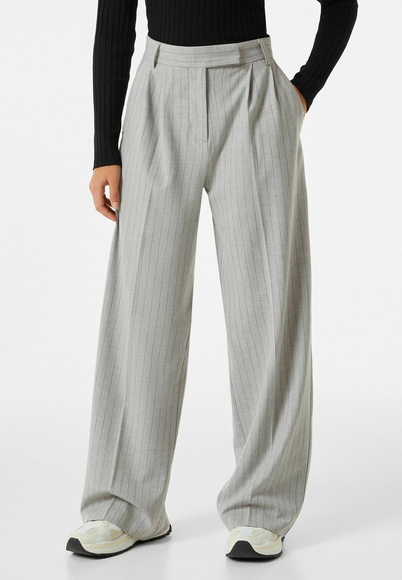 Femme portant un pantalon large gris clair à fines rayures et des baskets blanches, les mains dans les poches, montrée de la taille aux pieds.