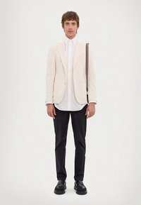 Férfi fehér blézerben és ingben, fekete nadrágban, fekete cipőben, egy csíkos pántú válltáskával, fehér háttér előtt áll.