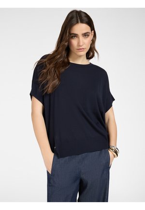 Mujer con cabello largo y castaño, vistiendo una blusa suelta de color azul marino oscuro y pantalones grises, de pie con las manos en los bolsillos contra un fondo blanco.