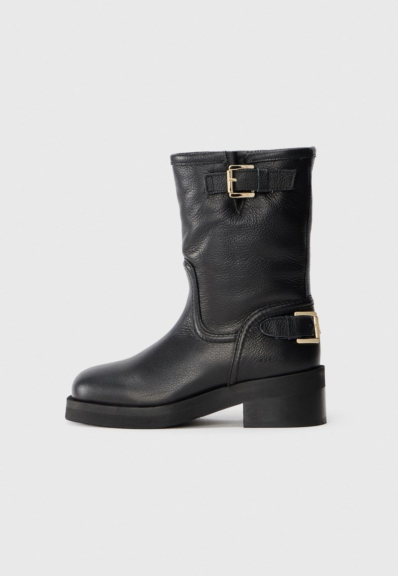 Botte en cuir noir mi-mollet avec deux lanières à boucle dorée, bout rond, talon bas et épais, finition texturée sur fond uni.
