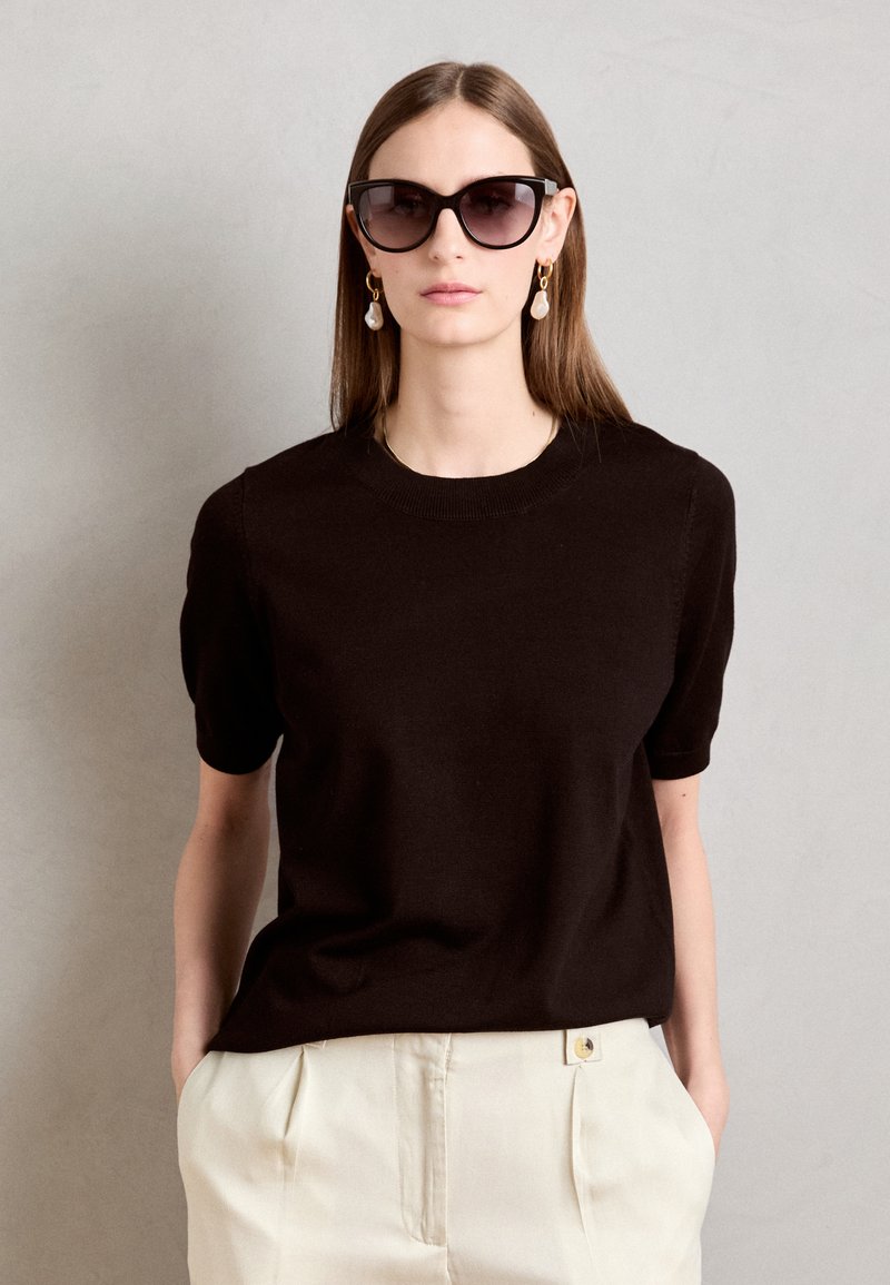 Pulover tricotat negru cu mânecă scurtă și guler rotund, asortat cu pantaloni plisati crem. Model purtând ochelari de soare supradimensionați negri și cercei lungi.