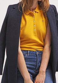 Gul pikétröja med krage och knappdetaljer, lagd under en mörk marinblå kappa, tillsammans med blå denimjeans.