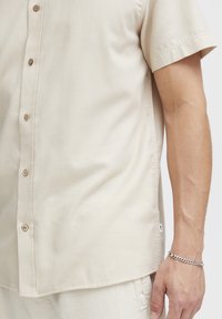 Beige kortom shirt met knopen in een zachte stof, voorzien van houten knopen, een gebogen zoom en een klein logotag aan de zijkant.