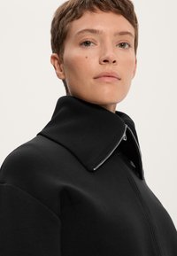 Musta vetoketjullinen takki, jossa on leveä ribbauskaulus ja teksturoitu kangas. Ominaisuuksiin kuuluu näkyvä ompelu ja tyylikäs, minimalinen muotoilu.