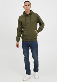 Olive green hoodie with drawstrings, front pocket, and ribbed cuffs; paired with blue denim jeans and white sneakers.