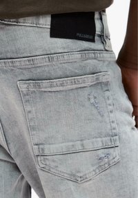 Svijetlo sivi traperice s vidljivim stražnjim džepom i crnom Pull&Bear etiketom blizu struka na osobi koja nosi tamnu majicu.