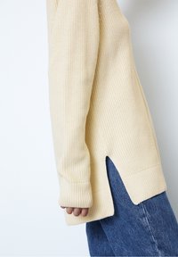 Pull en tricot couleur crème avec une texture côtelée, fentes latérales et manches longues, associé à un jean en denim bleu.