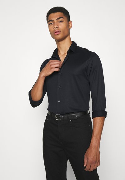 Camicia nera a maniche lunghe con bottoni e polsini arrotolati, texture liscia e colletto. Indossata con pantaloni neri e una cintura sottile.
