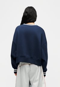 Γυναίκα με μακριά μαύρα μαλλιά φοράει υπερμεγέθη φούτερ σε ναυτικό μπλε και ανοιχτό γκρι παντελόνι με πορτοκαλί ρίγα, στέκεται γυρισμένη πλάτη σε απλό φόντο.