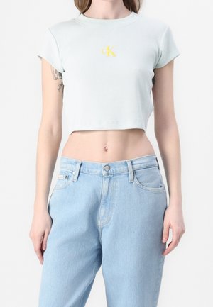 Kobieta w jasnoniebieskim krótko skrojonym T-shircie z żółtym logo "ck" i jasnoniebieskich, wysokich jeansach, stojąca na białym tle.