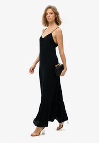 Sort, ærmeløs maxikjole med flæset nederdel, lavet af glat stof, sat sammen med en lille vævet clutch og sandaler med klare hæle.