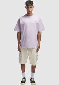 Lys lilla bomulls T-skjorte med oversized passform og rund hals, kombinert med kremfargede cargo-shorts med sidelommer og snøring. Svart sneakers.