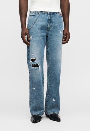 Svetlomodré rifle z opotrebovaného denimu so širokými nohavicami, ktoré majú veľké trhliny, jemné švy a kovový reťazový detail na páse.