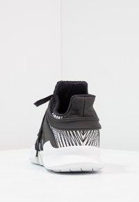 Svart och vit atletisk sko med mönstrad textilöverdel, svart hälstöd och tjock vit sula sedd från baksidan på en vit yta.