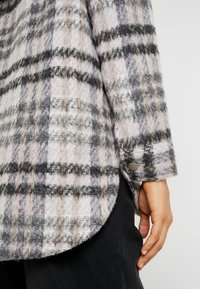 Wool blend shirt jacket in a grey, black, and cream plaid pattern; rounded hem; featuring buttoned cuffs.