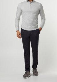 Hombre vestido con una camiseta de polo de manga larga gris claro, pantalones ajustados negros y zapatillas de ante grises, de pie con una mano en el bolsillo.