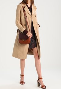 Manteau beige à double boutonnage, associé à un blouse violet foncé, une jupe à motifs, un sac bandoulière marron et des sandales à talons marron.