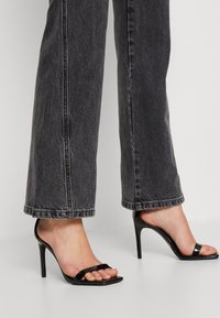 Sandalias de tacón alto de charol negro con punta cuadrada y dos tiras horizontales, combinadas con jeans de mezclilla oscura.
