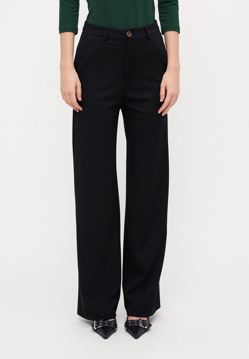 Femme portant un pantalon noir à jambes larges, un haut vert à manches longues et des chaussures noires pointues avec boucles argentées.