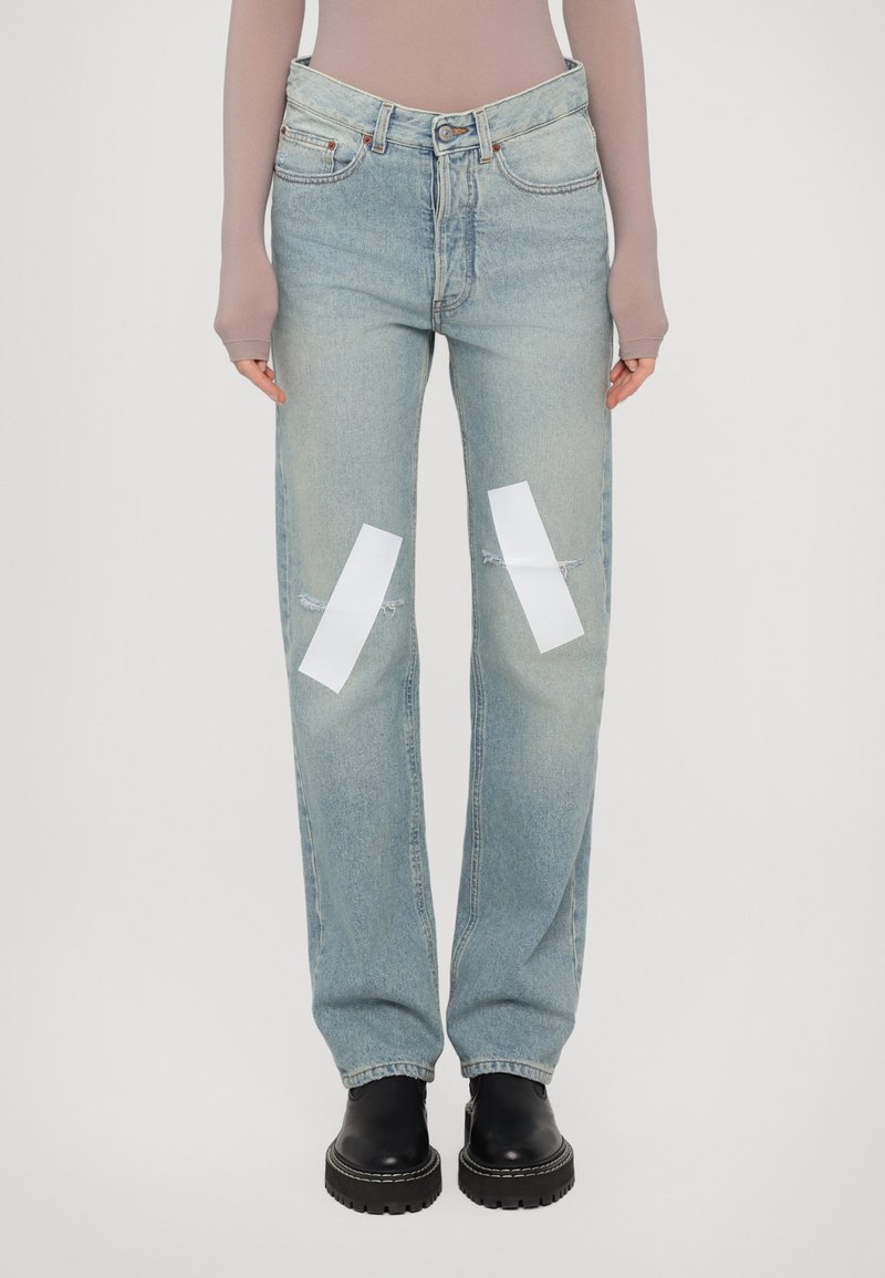 Helle blaue Denim-Jeans mit lockerer Passform, die mit distressierten Patches und weißen Bandakzenten an den Knien versehen sind. Schwarze klobige Stiefel runden den Look ab.