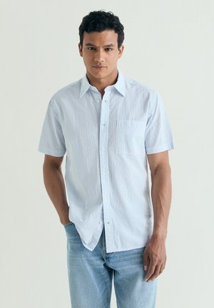 Hombre con camisa de manga corta color azul claro con finas rayas blancas, de pie con una mano en el bolsillo frente a un fondo liso.