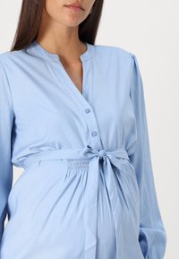 Světle modré košilové šaty s výstřihem do "V", dlouhými rukávy a zavázaným pasem. Hladká tkanina s nařasením v oblasti pasu.