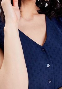 Navy sininen, teksturoitu paita, jossa on syvä V-kaula-aukko, pieniä kankaasta päällystettyjä nappia edessä ja sileä, pehmeä viimeistely.